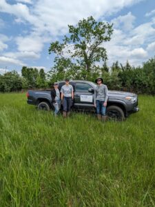 A Mad Scientist Associates, LLC truck with three people standing in front of it. The truck is parked in the grassy area near our in progress wetland area. 